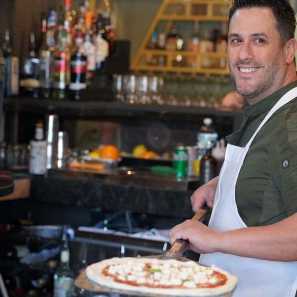 Smiling chef in a white apron holding a pizza peel with an uncooked pizza, bar in background.