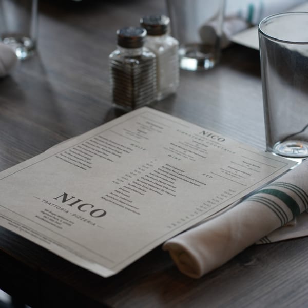 A Nico Trattoria Pizzeria menu, salt and pepper shakers, and a glass on a wooden table.