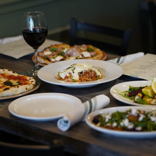 A table laden with various Italian dishes, including pizzas, pasta, and salads, with a glass of red wine.