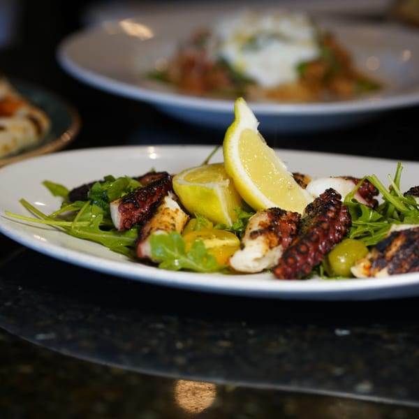 Grilled octopus salad with arugula, cherry tomatoes, olives, and lemon wedges on a white plate.